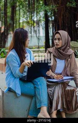 Un paio di donne indonesiane discutono candidamente di incarichi scolastici che trasportano computer portatili, libri e appunti nel parco del campus. studentesse da educare Foto Stock