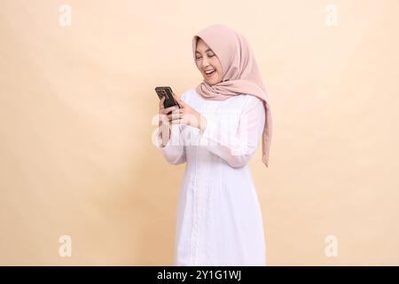 Allegra donna musulmana asiatica che indossa un hijab gestisce un gadget per cellulari con entrambe le mani. Stile di vita, tecnologia e concetto di promozione Foto Stock