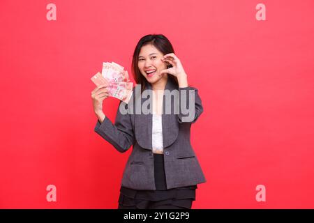 L'espressione di una donna indonesiana felice che tiene in mano una carta di credito, debito e denaro, mentre mostra amore per la macchina fotografica che indossa una giacca grigia e sci Foto Stock