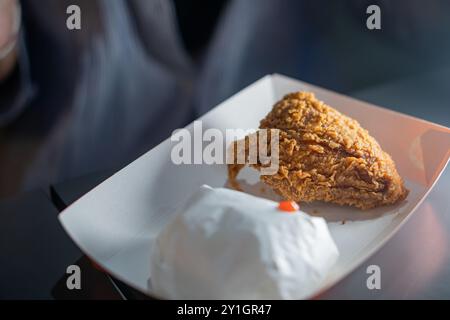 Un pezzo di pollo fritto croccante su un vassoio bianco, accompagnato da un piccolo contenitore bianco, eventualmente contenente un contorno o una salsa. Lo sfondo è B. Foto Stock