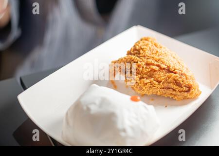 Un pezzo di pollo fritto croccante su un vassoio bianco, accompagnato da un piccolo contenitore bianco, eventualmente contenente un contorno o una salsa. Lo sfondo è B. Foto Stock