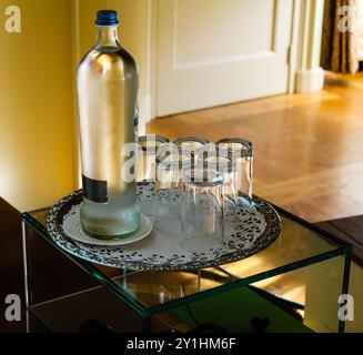 Una bottiglia d'acqua in vetro posta su un vassoio decorativo con sei bicchieri vuoti. L'ambiente e' interno, caratterizzato da pavimento in legno e illuminazione soffusa. Foto Stock