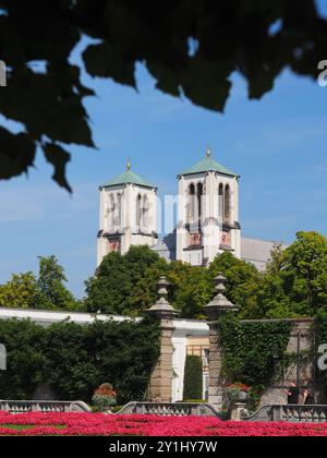 Salisburgo, Austria - 31 agosto 2024: Le torri gemelle della chiesa, con una splendida vegetazione in primo piano, si estendono verso un cielo blu luminoso, che mostra l'eleg Foto Stock