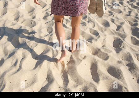 Donna con un prendisole vista l'anca da dietro mentre cammina a piedi nudi su una spiaggia sabbiosa, tenendo le scarpe in una mano. Foto Stock