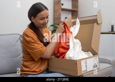 Una donna sorridente con capelli scuri toglie abiti luminosi ordinati dal negozio online dalla scatola di cartone Foto Stock