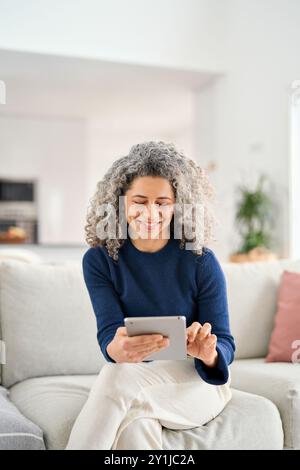 Donna di mezza età felice che utilizza un tablet digitale e si rilassa sul divano di casa. Foto Stock