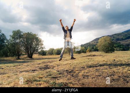 Giovane energico che salta con le braccia sollevate nella vittoria poggia su un cielo spettacolare e sullo sfondo di montagne. Concetto di realizzazione, libertà e connessione Foto Stock