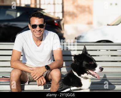 Un uomo siede su una panchina con il suo cane. Quell'uomo sorride e indossa gli occhiali da sole. Il cane è seduto accanto a lui ed è felice. La scena è rilassata Foto Stock