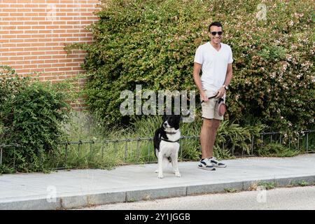 Un uomo sta camminando il suo cane su un marciapiede. Il cane è bianco e nero. L'uomo indossa una camicia bianca e pantaloncini Foto Stock