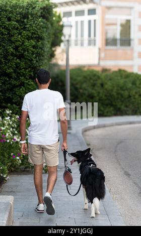 Un uomo sta camminando il suo cane su un marciapiede. Il cane è bianco e nero e indossa un guinzaglio. La scena è tranquilla e calma, con l'uomo Foto Stock