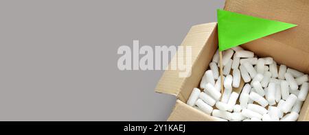 Scatola di imballaggio in cartone aperta con chip di imballaggio in polistirene bianco all'interno. Indicatore di carta di colore verde per nota, avviso, testo. Imballaggio, spedizione, shopping Foto Stock