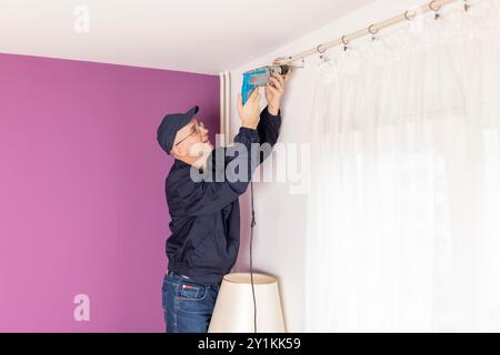 Un uomo che lavora, si occupa di ristrutturazioni domestiche, costruzioni o falegnameria. Un esperto artigiano maturo con cacciavite elettrico lavora con piacere Foto Stock