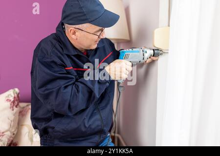 Un uomo che lavora, si occupa di ristrutturazioni domestiche, costruzioni o falegnameria. Un esperto artigiano maturo con cacciavite elettrico lavora con piacere Foto Stock