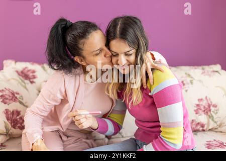 Figlia felice che mostra il test di gravidanza a sua madre sul divano. Due facce di donne irradiano felicità ed eccitazione mentre guardano la gravidanza Foto Stock