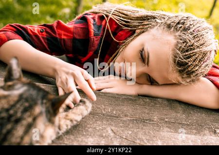 Una giovane donna con trecce bionde accarezza il suo gatto su un portico di legno, mostrando amore e amicizia in un sereno momento all'aperto Foto Stock