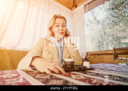 Telefonata donna Coffee - donna sorridente seduta ad un tavolo con una tazza di caffè, che parla al telefono in un caffè o in un'area salotto all'aperto. Foto Stock