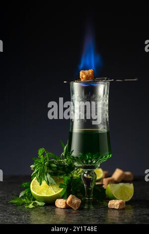 Bruciare lo zucchero su un bicchiere con assenzio. Preparazione della bevanda. Absinthe con ingredienti su sfondo nero. Foto Stock