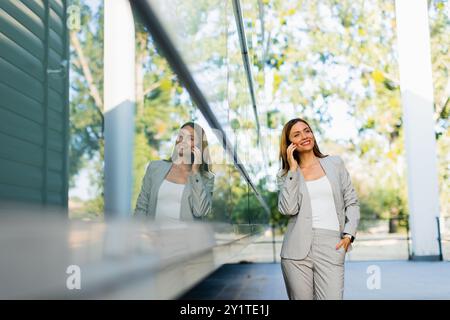 Una donna professionista cammina con facilità, sorridendo mentre si concentra sul telefono, mescolando lo stile di vita moderno con la tranquillità della natura sullo sfondo Foto Stock