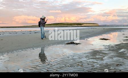 Donna escursionista con zaino in spalla che gode del paesaggio costiero del tramonto sulla spiaggia sabbiosa di Silverstrand, Galway, Irlanda, avventura e stile di vita, persone nella natura Foto Stock