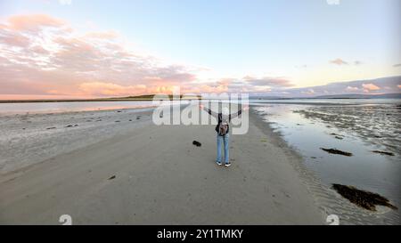 Donna escursionista con zaino in spalla che gode del paesaggio costiero del tramonto sulla spiaggia sabbiosa di Silverstrand, Galway, Irlanda, avventura e stile di vita, persone nella natura Foto Stock