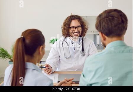 Medico di famiglia sorridente che siede alla scrivania e parla con una coppia in clinica medica. Foto Stock