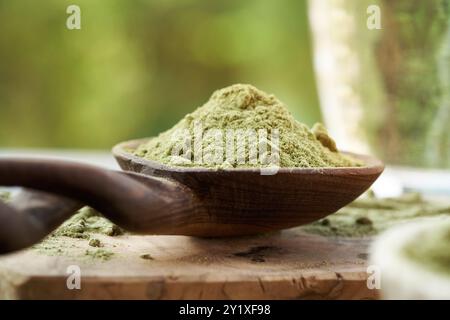 Erba d'orzo in polvere su un cucchiaio di legno all'aperto in natura, con un bicchiere d'acqua sullo sfondo Foto Stock