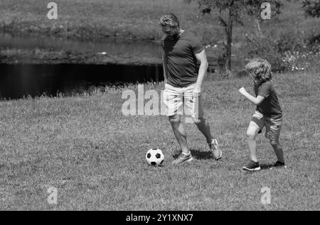 Padre con figlio che gioca a calcio sul campo. Padre e figlio giocano a calcio insieme nel parco estivo. Il figlio eccitatamente calcia la palla verso il padre Foto Stock