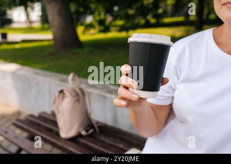 Primo piano di una donna irriconoscibile che si gode del tempo libero all'aperto sorseggiando un caffè seduto sulla panchina del parco cittadino. Donna felice che riposa da sola Foto Stock
