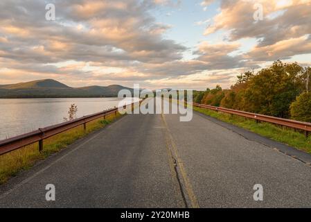 Strada vuota che attraversa una diga in un paesaggio montano boscoso sotto il cielo spettacolare in autunno. Foto Stock