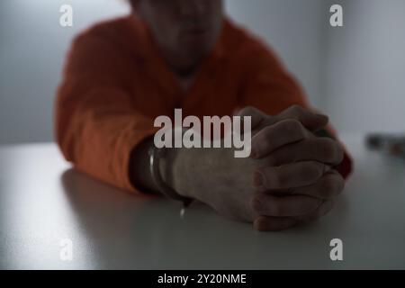 Detenuto che indossa una tuta arancione brillante seduta con le mani ammanettate sul tavolo nella sala degli interrogatori. Il volto sfocato e la luce soffusa aumentano la gravità della scena Foto Stock