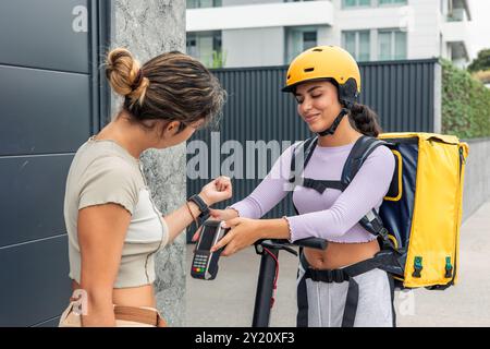 La donna che effettua il servizio di consegna in latino, dotata di zaino e casco giallo, fornisce un lettore di carte mentre un cliente effettua il pagamento utilizzando uno smartwatch. mostra Foto Stock