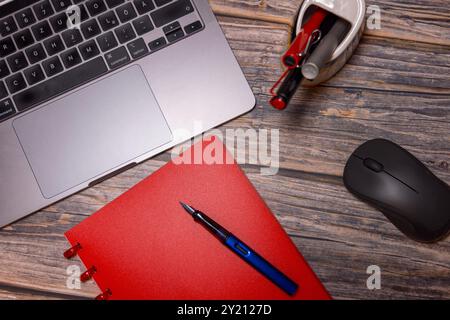 Una composizione piatta con un notebook rosso, una penna stilografica, un computer portatile e un mouse su una scrivania rustica in legno, che rappresenta l'essenziale per lo spazio di lavoro moderno Foto Stock