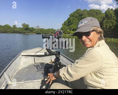 Ecoturismo femminile guardando la macchina fotografica con un treppiede e macchina fotografica su una piccola barca tour su un fiume in Pantanal Wetland, alberi verdi sullo sfondo Foto Stock