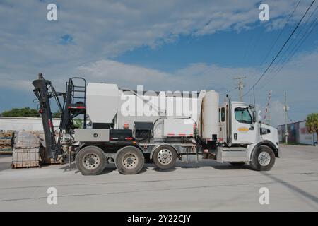 Vista laterale di un grande camion di cemento bianco nel tardo pomeriggio con cielo blu e nuvole bianche sul retro. Binari ferroviari di fronte alla strada polverosa con edificio Foto Stock