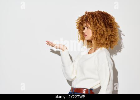 Donna dai capelli ricci mostra un gesto espressivo della mano su uno sfondo bianco minimalista, che enfatizza la sicurezza e lo stile del look alla moda Foto Stock