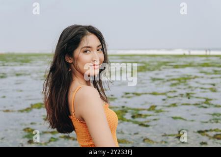 La giovane donna asiatica chic che indossa un abito arancione guarda la macchina fotografica dalla sua spalla con l'oceano sullo sfondo. Foto Stock