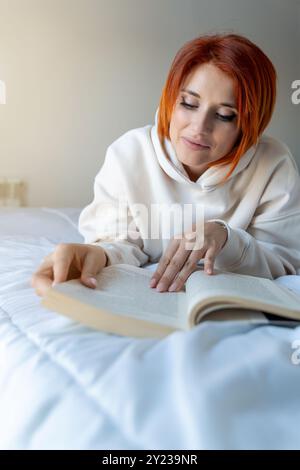 Una donna dai capelli rossi in una felpa con cappuccio beige giace comodamente su un letto, leggendo un libro. Foto Stock