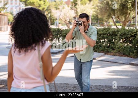 Un uomo sorride mentre scatta una foto di una donna che posa giocosamente in un parco soleggiato. Godono di un momento di luce circondato da verde e luce naturale. Foto Stock