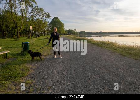 La donna sta facendo una tranquilla passeggiata serale con il suo cane lungo un sentiero vicino a un lago sereno, il sole tramonta proiettando un caldo bagliore sull'acqua Foto Stock