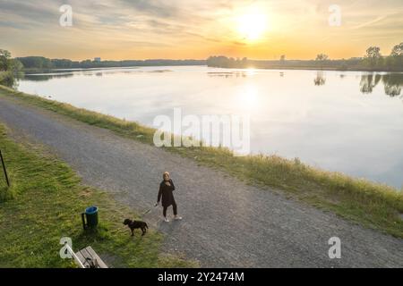 La donna sta facendo una tranquilla passeggiata serale con il suo cane lungo un sentiero vicino a un lago sereno, il sole tramonta proiettando un caldo bagliore sull'acqua Foto Stock