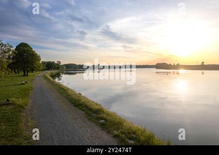 Il tranquillo sentiero di ghiaia si snoda lungo la tranquilla riva di un lago al tramonto, con una luce dorata che si riflette nelle acque calme, creando una scena tranquilla, po R. Foto Stock