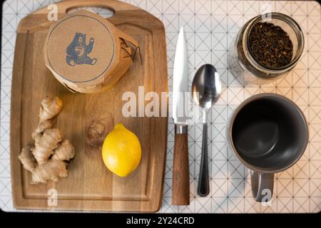 Preparazione del tè con miele, zenzero, limone e foglie di tè sciolte, disposte su un asse di legno con una tazza, un coltello e un cucchiaio. Foto Stock