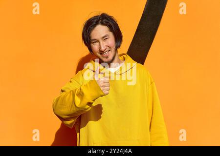 Un ragazzo asiatico con una felpa gialla si erge e mostra un gesto super, un Foto Stock