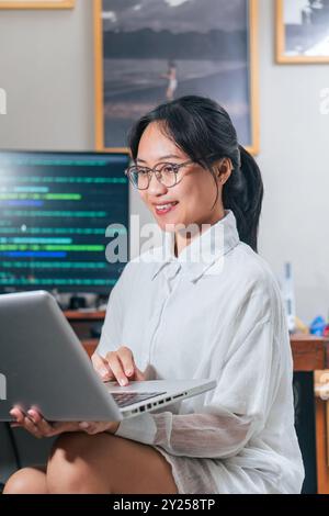 Una giovane donna con gli occhiali che lavora su un notebook in un ufficio moderno. Appare concentrata e impegnata, con uno sfondo con foto incorniciate Foto Stock