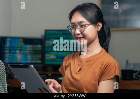 Una giovane donna con gli occhiali sta usando un tablet in un ufficio moderno. Sembra concentrata e impegnata, con più schermi di computer che visualizzano da Foto Stock