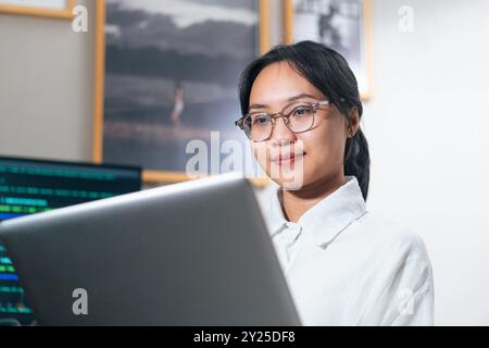 Una giovane donna con gli occhiali che lavora su un notebook in un ufficio moderno. Appare concentrata e impegnata, con uno sfondo con foto incorniciate Foto Stock