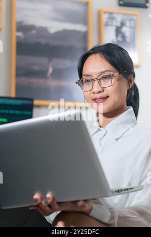 Una giovane donna con gli occhiali che lavora su un notebook in un ufficio moderno. Appare concentrata e impegnata, con uno sfondo con foto incorniciate Foto Stock