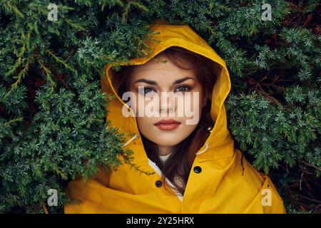 Donna con impermeabile giallo che si nasconde dietro alberi sempreverdi, sentendosi protetta e isolata in natura in una giornata di pioggia Foto Stock