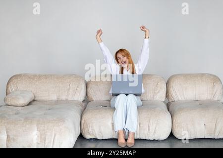 Excited donna usando il telefono e il computer portatile seduto su un divano nel soggiorno di notte a casa Foto Stock