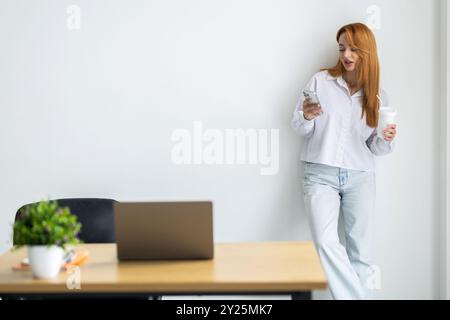 Bella giovane donna d'affari intelligente sorridente in piedi in ufficio con un gruppo di colleghi sullo sfondo, utilizzando il telefono cellulare, tenendo in mano il co-takeaway Foto Stock
